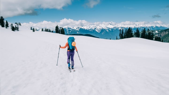 Backcountry skier in purple leggings, orange top, and blue backpack ascends a snow-covered hill in Garibaldi Park near Squamish