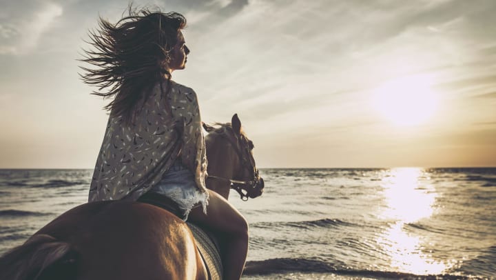Back view of a woman riding a horse on the beach at sunset.
