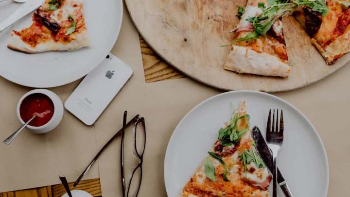 Flat-lay shot of a table set for a casual meal, featuring several slices of thin-crust pizza topped with arugula and prosciutto. The slices are on two white plates and a round wooden serving board. Also visible on the brown table surface are an iPhone, dark-framed eyeglasses, and small bowls of red and white dipping sauces.