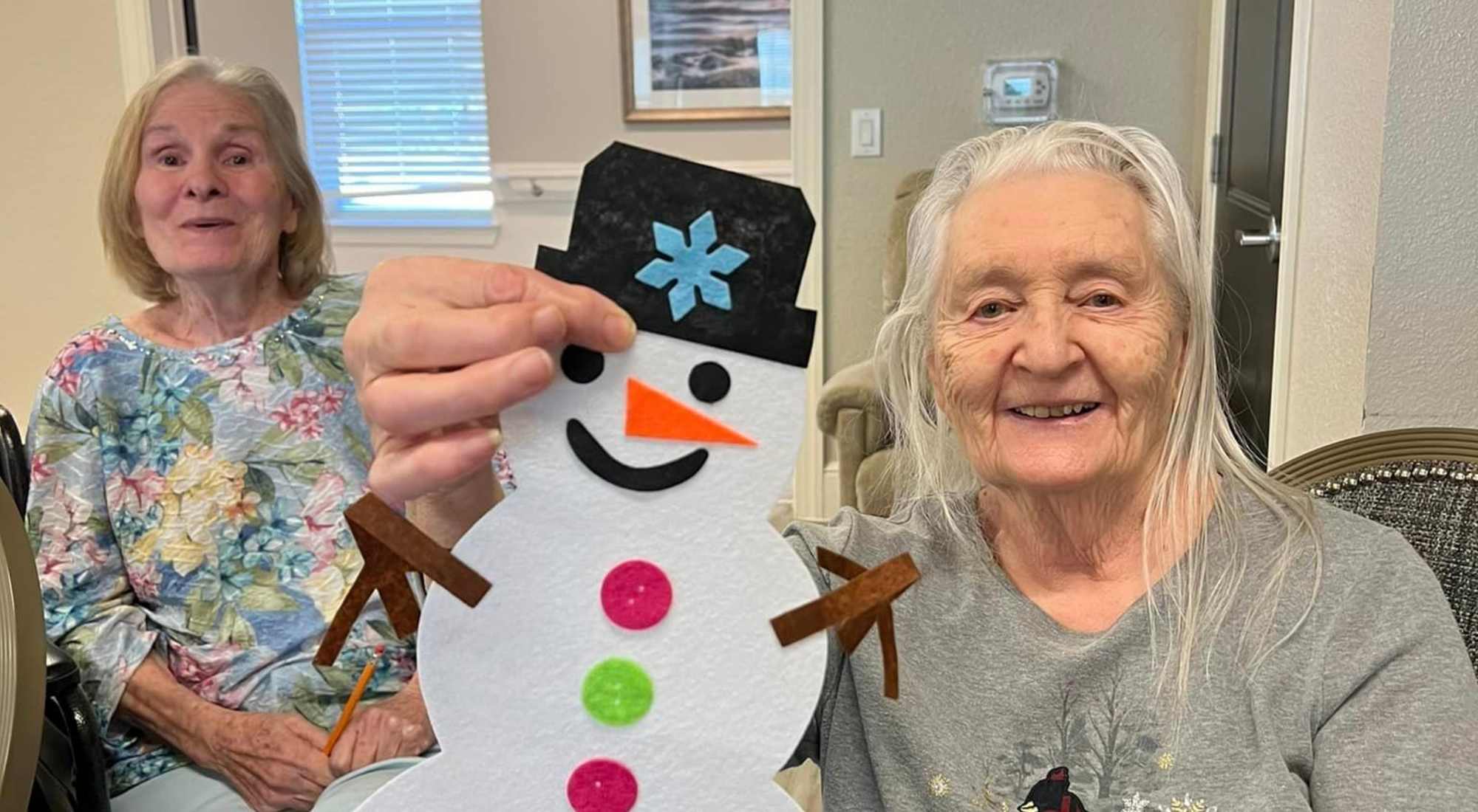 Two smiling elderly women showing off a cheerful handmade snowman craft.