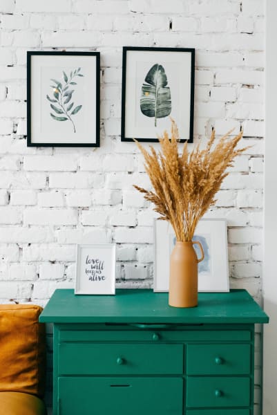 A painted green dresser with a painted vase full of wheat grains in front of a painted white brick wall with frame photos hanging. 