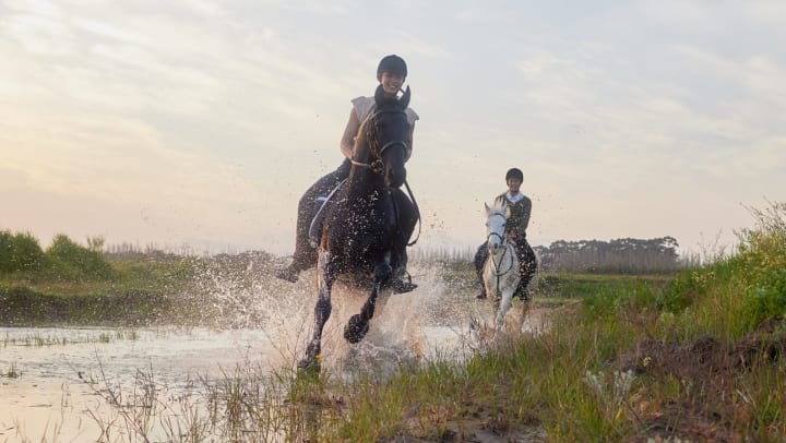 Two women riding horses through a puddle of water.