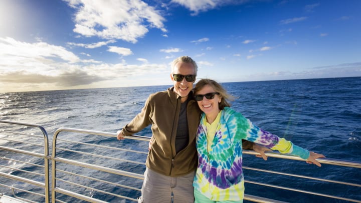 A senior man and woman leaning against the rails on a ship smiling with the ocean in the background. 