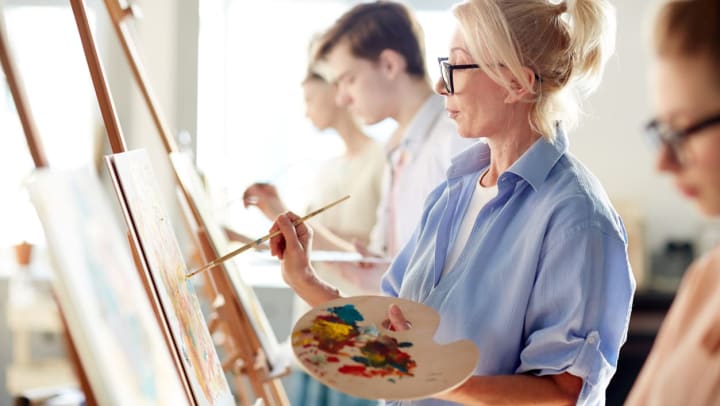 A woman with blonde hair and a blue button up shirt paints on a canvas | painting lessons in Keller