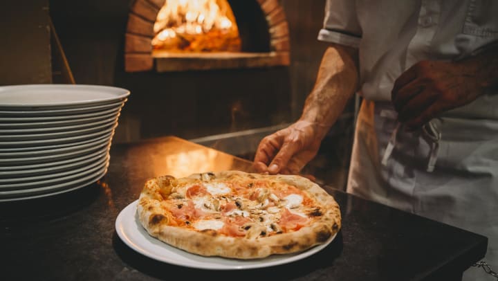 Wood fired pizza on a plate with cheese and prosciutto with pizza oven and chef in the background | best pizza around Carollton