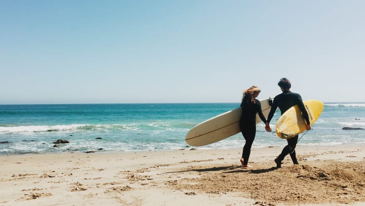 Couple holding hands on the beach, each with a surfboard under an arm.