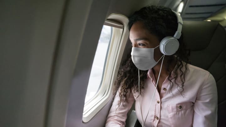 Young woman wearing a mask and headphones sitting in an airplane and looking out the window