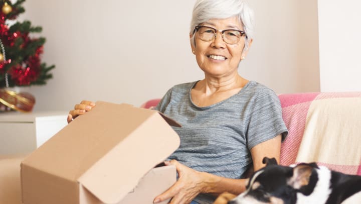 Senior woman sitting on a couch next to a sleeping dog, opening a box, with a Christmas tree in the background. 