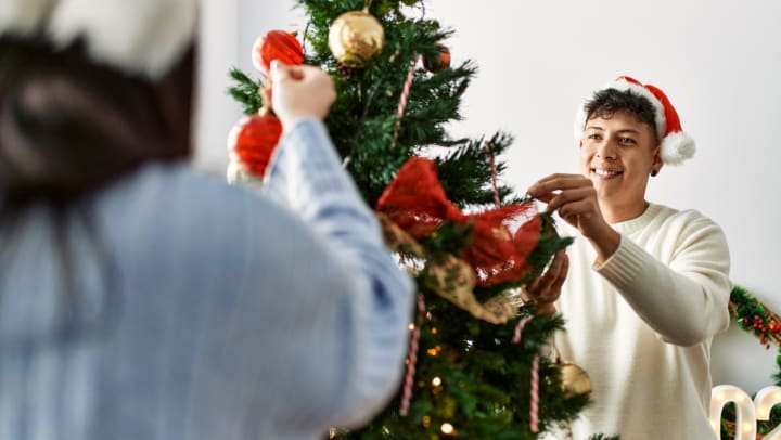 A young couple smiling and wearing Santa hats while decorating a tree.