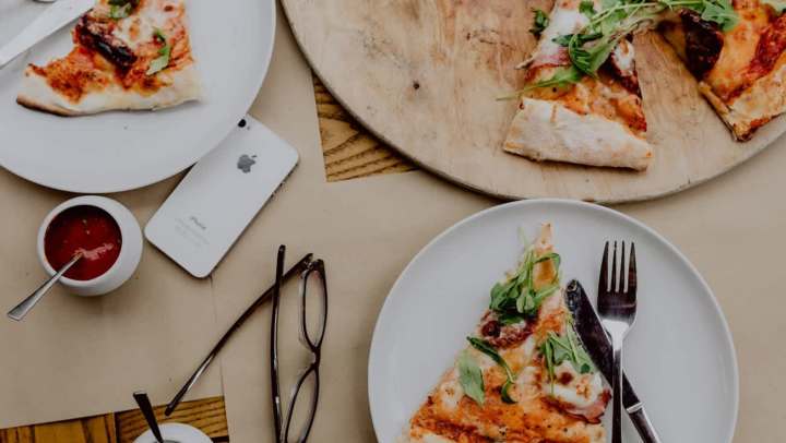 Flat-lay shot of a table set for a casual meal, featuring several slices of thin-crust pizza topped with arugula and prosciutto. The slices are on two white plates and a round wooden serving board. Also visible on the brown table surface are an iPhone, dark-framed eyeglasses, and small bowls of red and white dipping sauces.