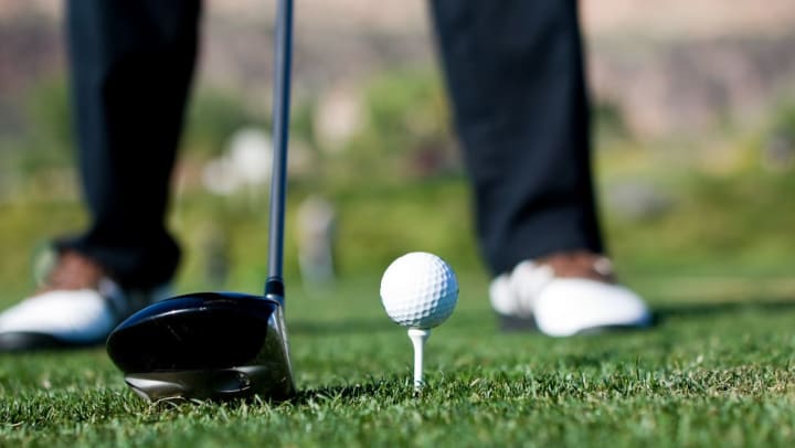 A golf ball sitting on top of a white tee, a driver at the ready and a pair of golfer