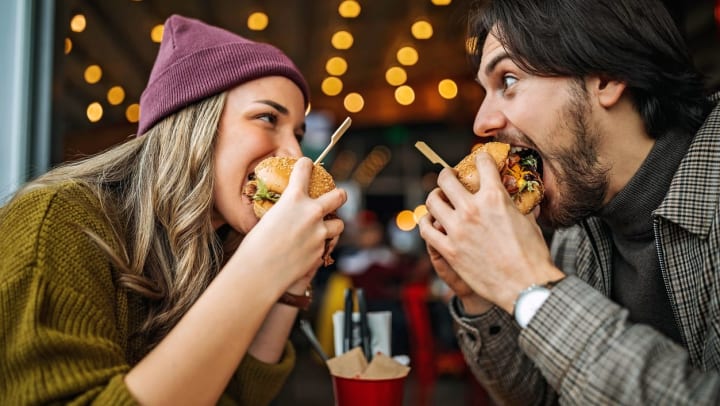 A man and woman excitedly look at each other across a table as they both bite into gourmet hamburgers