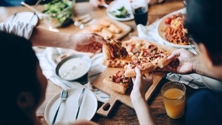  Overhead shot of a wooden table surrounded by diners. Among the white plates and napkins, people reach and pull slices from a centered wood platter containing a thick-crust pizza.