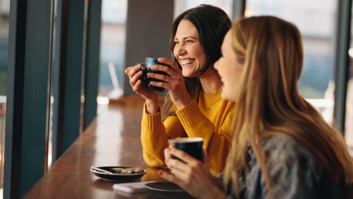 Two women laughing while enjoying cups of coffee by a window | coffee shops in Casper
