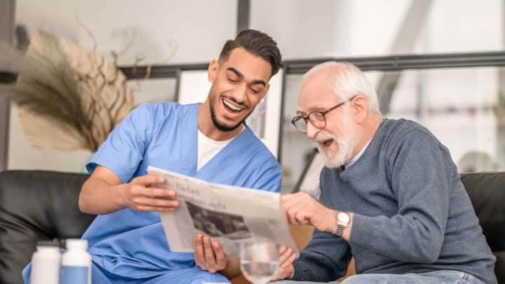 Older man and younger man in scrubs sitting on a couch and laughing at newspaper