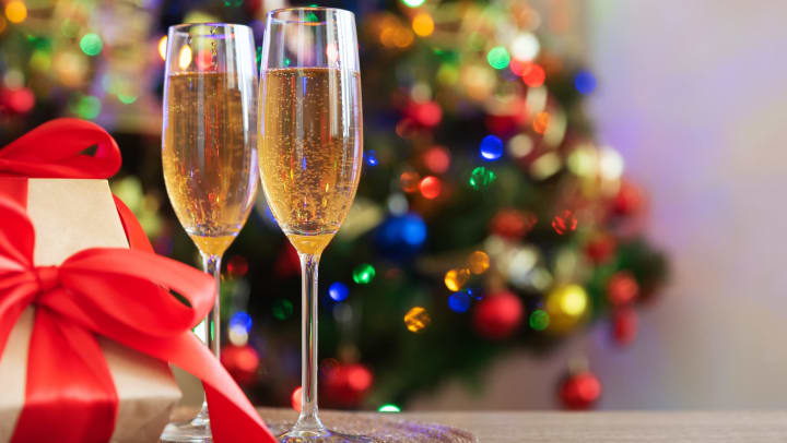 Champagne flutes full of a bubbly liquid next to a wrapped gift with a blurry lighted tree in the background. 