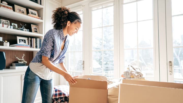 A woman smiling while packing up cardboard boxes in front of a sunny living room window