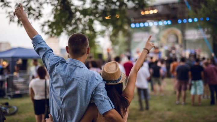 A couple each raises an arm in the air as they look toward the stage at an outdoor music festival. 