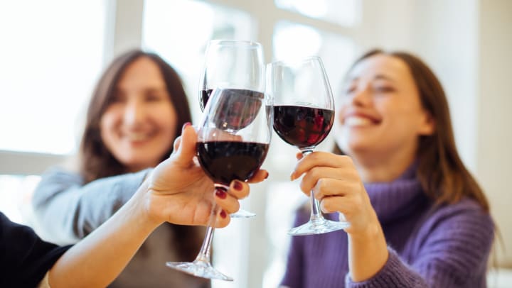 3 women touching wine glasses. The three glasses are in the foreground. Two of the three women can be seen in the background. 