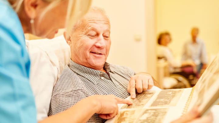 health care professional looking through old picture album with senior adult male reclined in bed in memory care facility