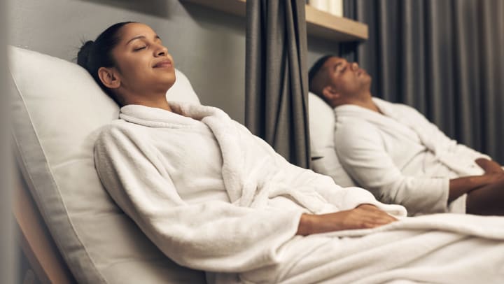 A man and a woman wearing white robes both relax on reclined chaises during a visit to a Brunswick spa.