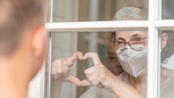 Elderly woman holding heart hands towards loved one outside of window