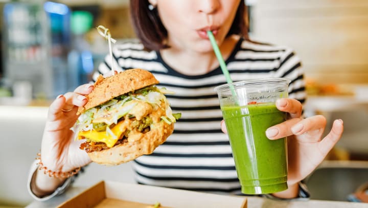 A woman holding a cup and drinking green juice from a straw while holding a sandwich in her other hand 