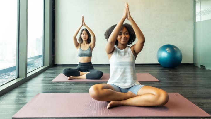 Two women sit on yoga mats in a hot yoga studio