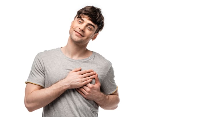 A smiling young man holding his hands over his heart while looking at the camera against a white backdrop.