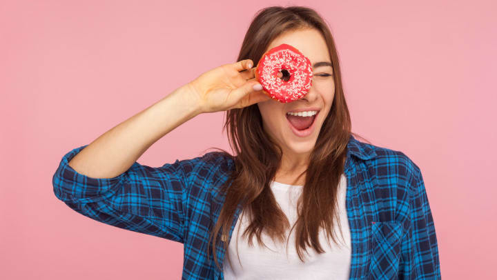 Woman looking through the hole in a pink frosted donut with white sprinkles standing in front of a pink background.
