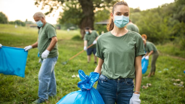 A woman in a mask and work gloves holds a large plastic bag of trash, while other volunteers in the background continue cleaning up trash from a natural area. 