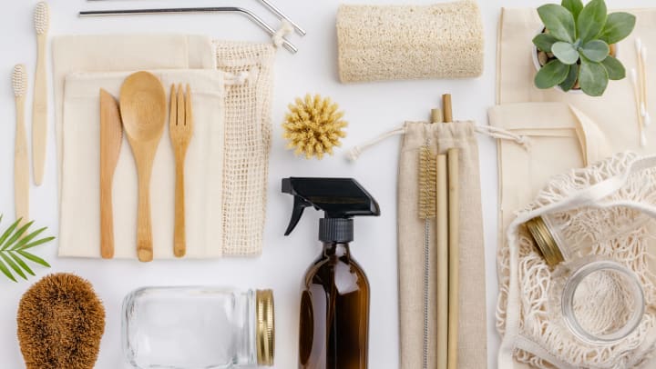 Natural color eco bags, reusable metal and bamboo straws, glass jars, wooden knives and forks, and zero waste cleaning and beauty products, against a white backdrop.