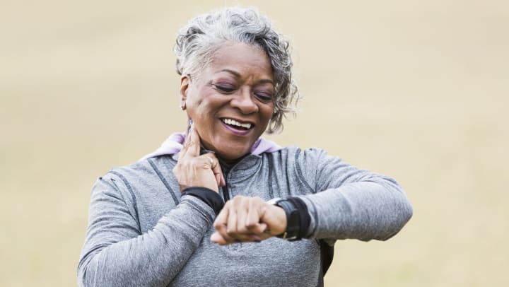 Smiling senior woman wearing athletic clothing checking her carotid pulse while walking outdoors.