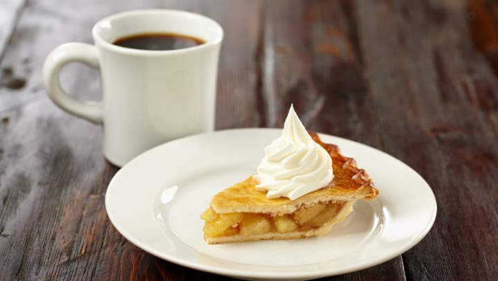 Cup of coffee next to a slice of apple pie with whipped cream on top.