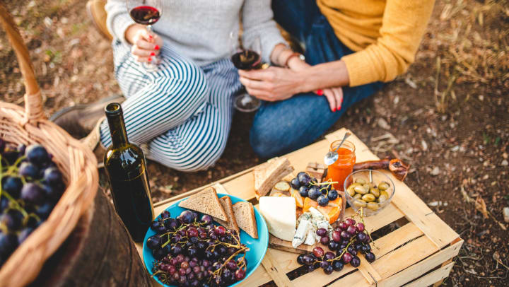Coupe sitting close together on the ground with wine glasses in their hands and grapes and cheese spread out on a wooden crate in front of them. 