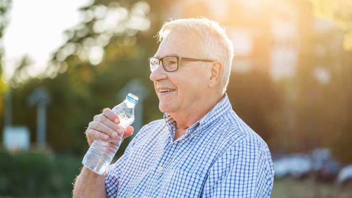 Older adult with glasses sitting on a park bench, drinking water in warm sunlight at {{location_name}} in {{location_city}}, {{location_state_name}}