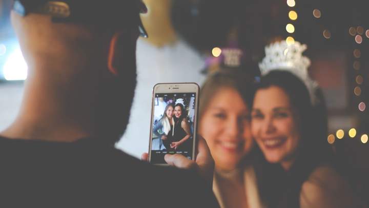 A man capturing a moment at a party, photographing two women smiling together.