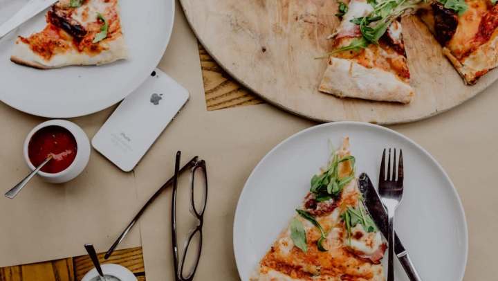 Flat-lay shot of a table set for a casual meal, featuring several slices of thin-crust pizza topped with arugula and prosciutto. The slices are on two white plates and a round wooden serving board. Also visible on the brown table surface are an iPhone, dark-framed eyeglasses, and small bowls of red and white dipping sauces.