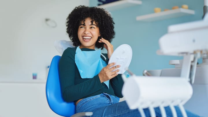 Woman smiling while sitting on dental exam chair and holding mirror | dental offices in Albuquerque