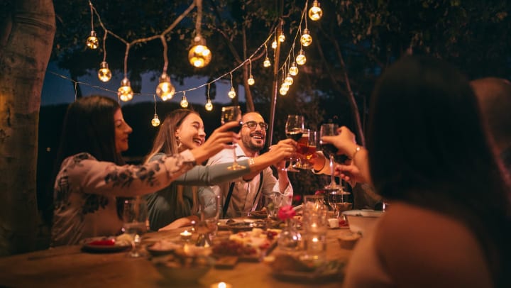 Smiling young people toasting around an evening dinner table lit by glowing string lights overhead.