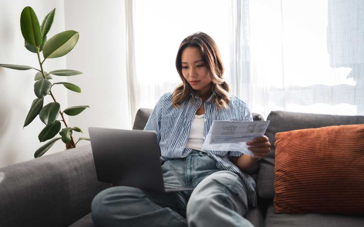 Woman sitting on couch using laptop, holding documents, with indoor plant nearby and natural light coming through windows at Carlton Arms North in Tampa, Florida