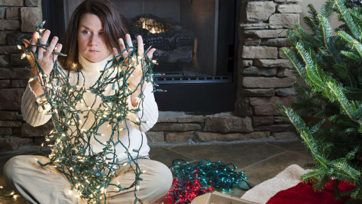 A young woman holds a tangle of string lights while sitting on the floor next to a holiday tree and boxes of ornaments.