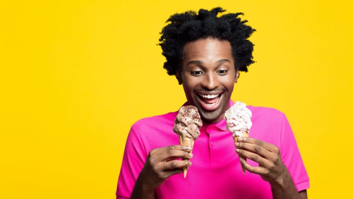 Man in a pink shirt looking at two ice cream cones with an open-mouth smile standing in front of a bright yellow background.