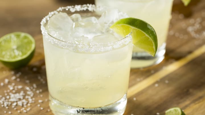 Margarita in a short glass with a salt rim and a lime on the rim with another out of focus just behind it. 
