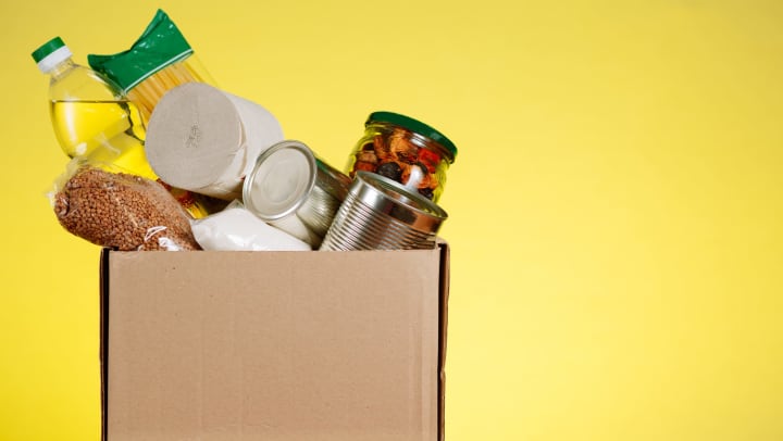 Cardboard box of various canned foods and household items against a yellow background