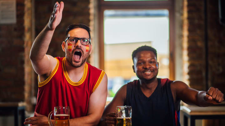 Two men in a bar cheering. One man has a basketball. They’re both wearing basketball jerseys