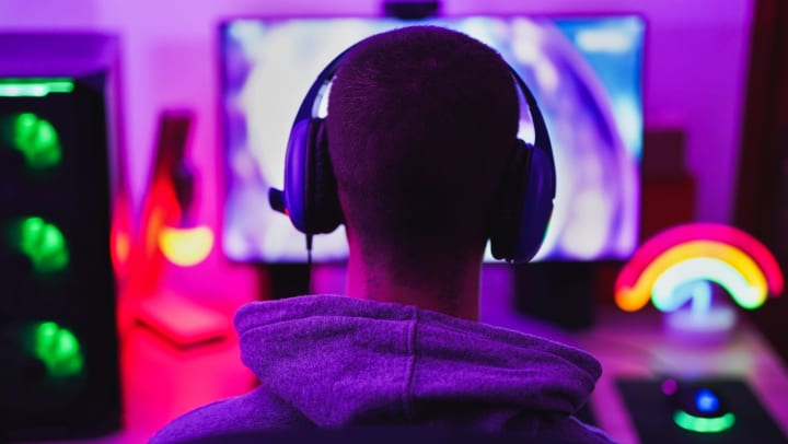 Person with blue headphones is facing a colorful computer screen with purple lighting. 