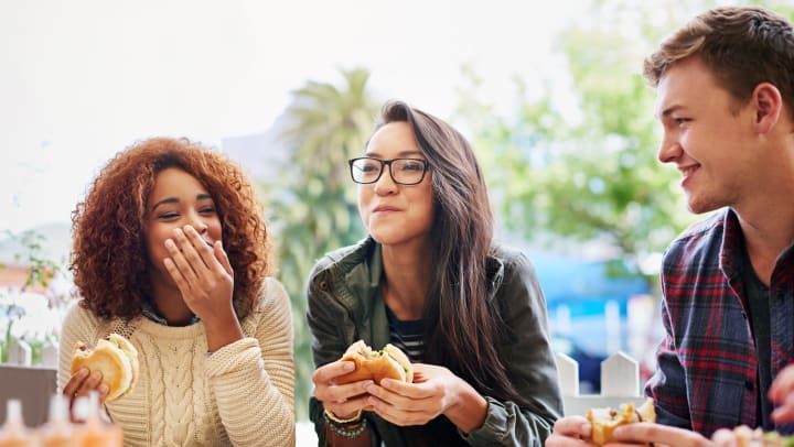 Three young people sitting outdoors with hamburgers in their hands while they smile.
