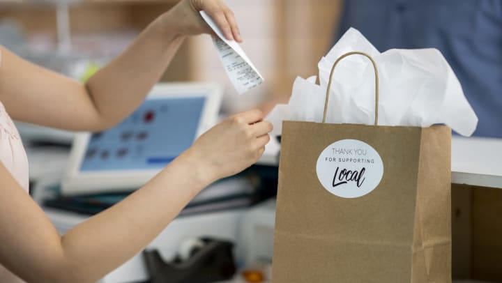 Retail worker placing receipt in a paper shopping bag with a "Thank You for Supporting Local" sticker on it.