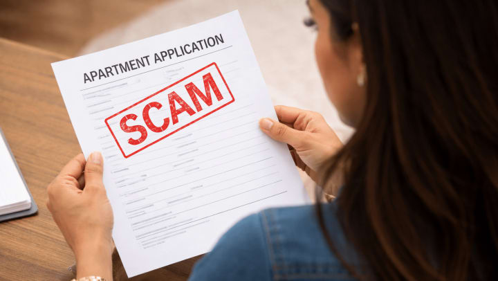 Over-the-shoulder view of a woman holding and reviewing an apartment application form stamped with the word “SCAM” in bold red letters.